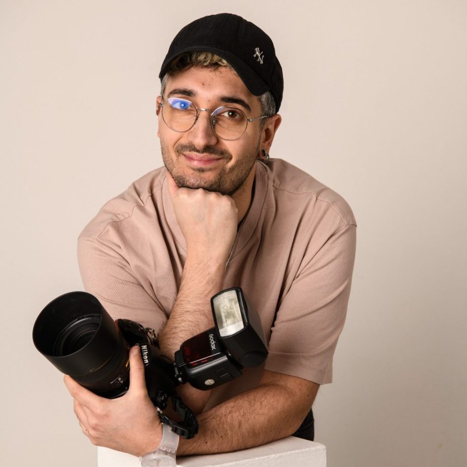 Uomo con telecamera su treppiede, indossa t-shirt rosa e cappellino, in un ambiente interno.