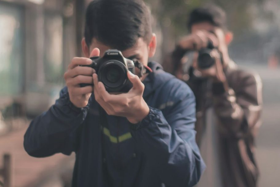 Due persone che scattano foto in una strada urbana, con alberi sullo sfondo.