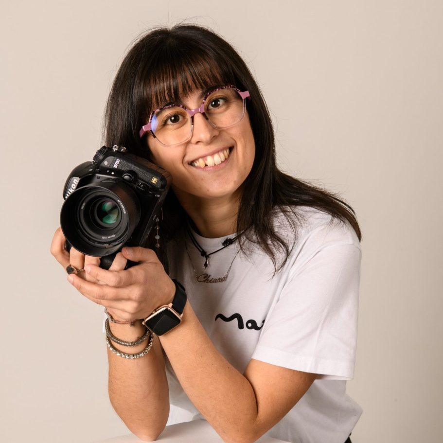 Giovane fotografa sorridente tiene una macchina fotografica, indossando occhiali e maglietta bianca.